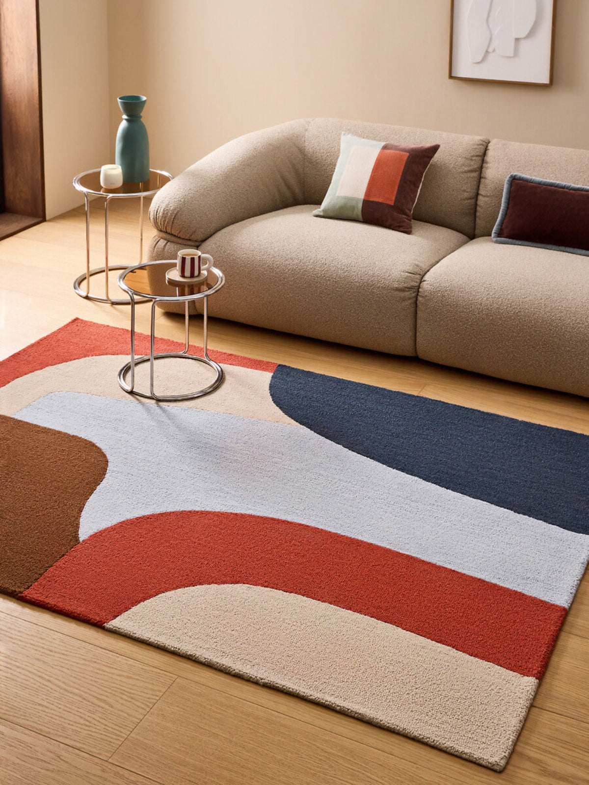 A modern abstract-pattern area rug in beige, rust red, navy blue, and brown tones, styled in a living room setting on a light wood floor in front of a beige sofa.