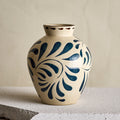 A ceramic vase with a cream-colored glaze and dark blue floral scroll patterns, displayed on a textured stone surface against a neutral beige background.