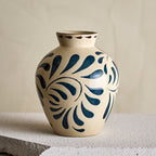 A ceramic vase with a cream-colored glaze and dark blue floral scroll patterns, displayed on a textured stone surface against a neutral beige background.