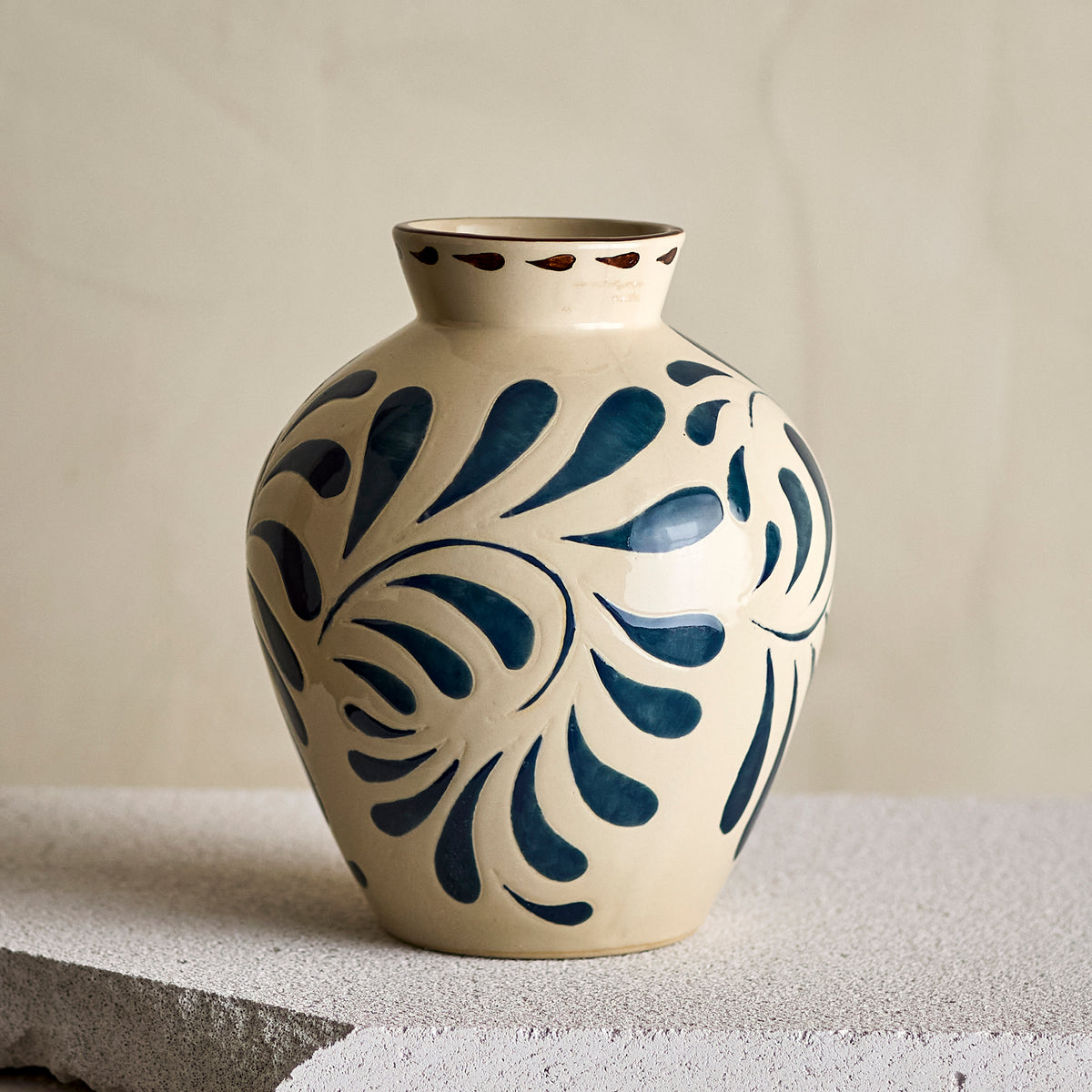 A ceramic vase with a cream-colored glaze and dark blue floral scroll patterns, displayed on a textured stone surface against a neutral beige background.