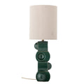 A modern table lamp with a cylindrical off-white fabric shade and a sculptural dark green ceramic base featuring circular and rectangular forms.