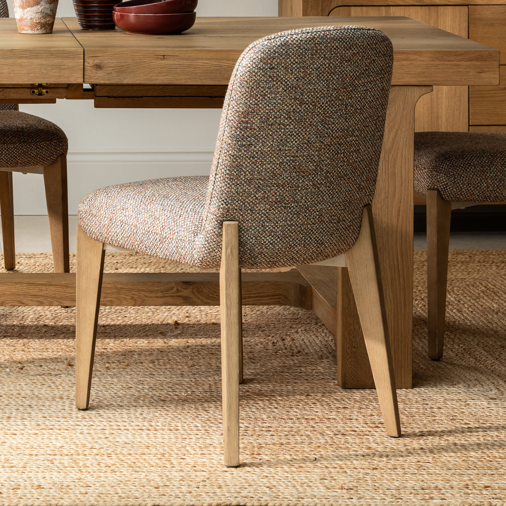 A close-up of a dining chair with a textured beige and brown herringbone upholstered seat and back, supported by light natural wood legs, positioned in front of a wooden dining table on a woven jute rug.