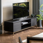 A black mango wood media unit with a gold-finish metal base, featuring a television on top and storage.