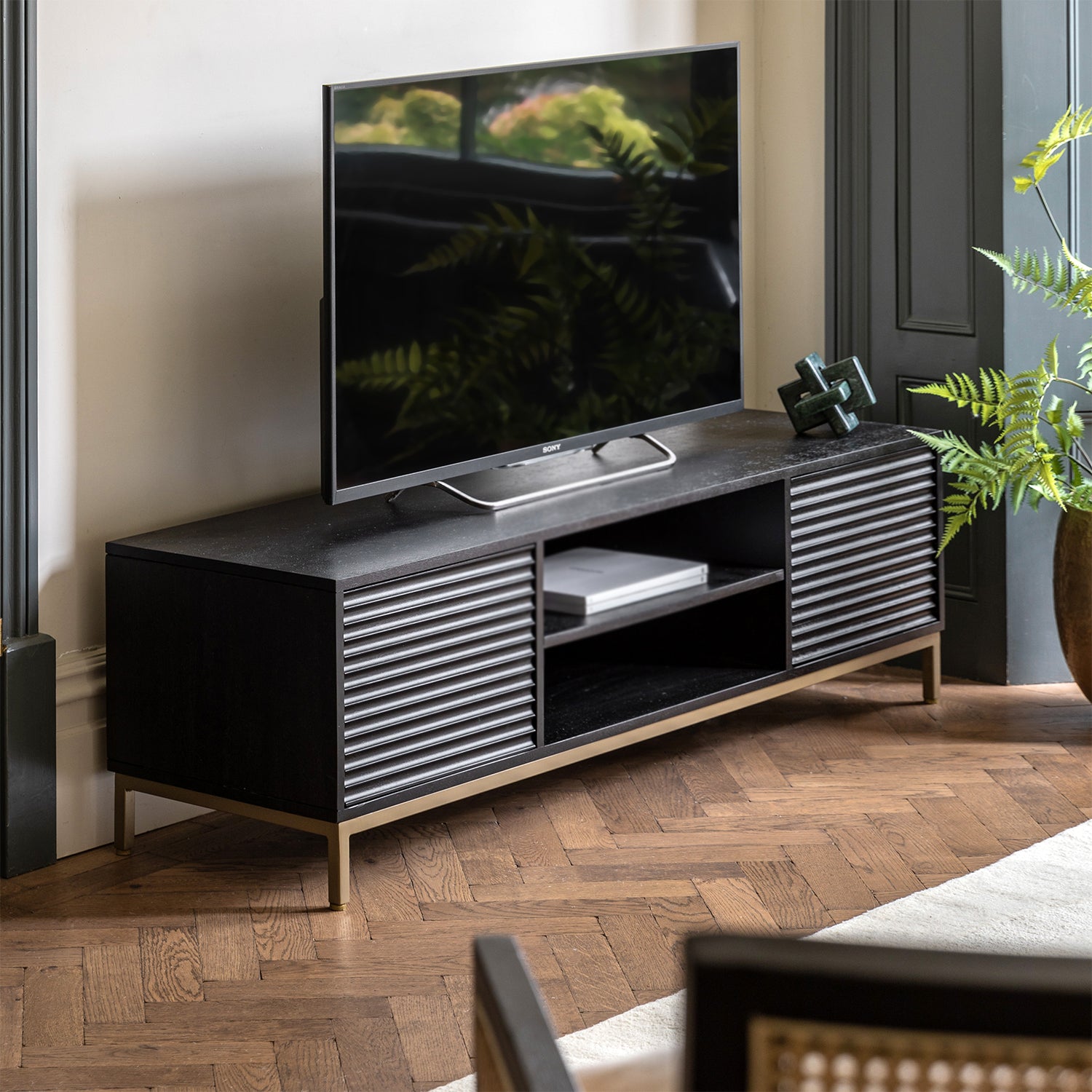 A black mango wood media unit with a gold-finish metal base, featuring a television on top and storage.
