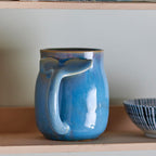 Blue ceramic mug with a handle on a wooden shelf with books and a glass jar.