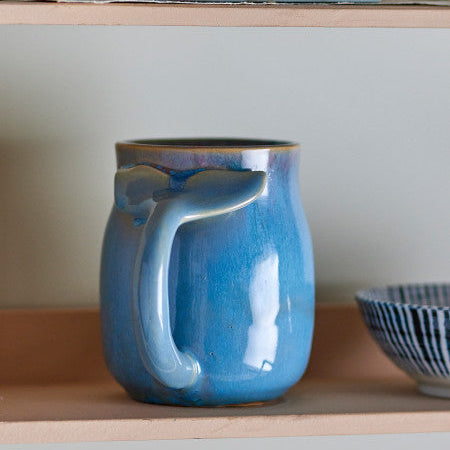 Blue ceramic mug with a handle on a wooden shelf with books and a glass jar.