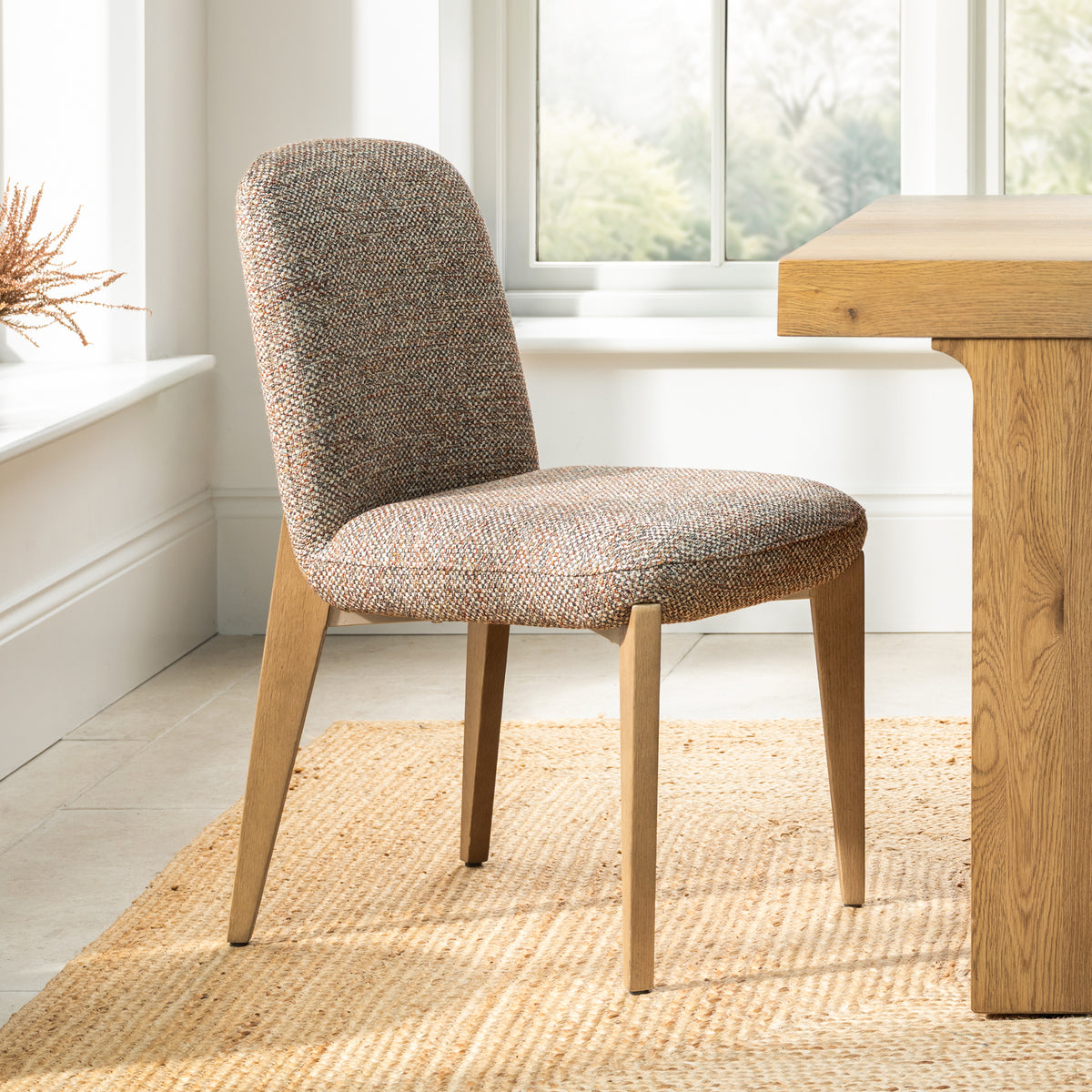 A modern upholstered dining chair with a textured brown and beige woven fabric seat and back, supported by four tapered light oak legs, shown in a bright room next to a wooden table.