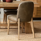 Taupe-brown leather upholstered seat dining chair, with light natural oak legs, shown in a dining room next to a wooden dining table.