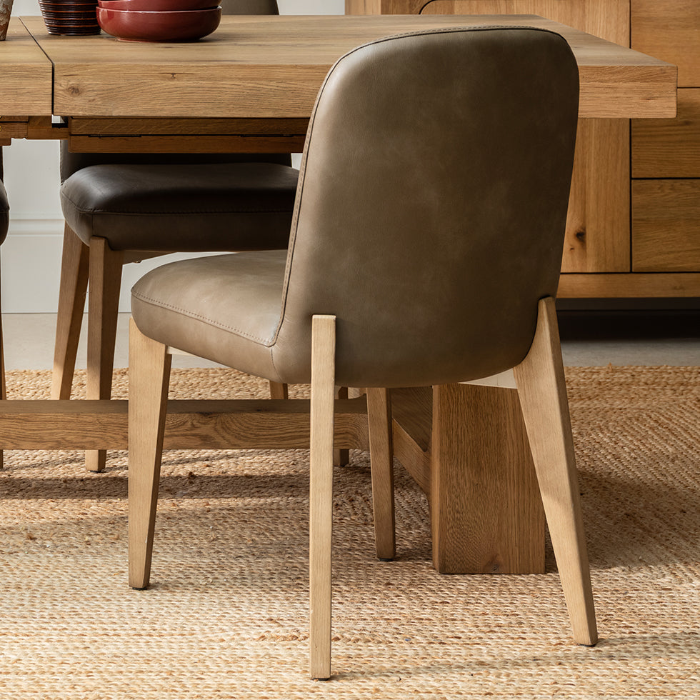 Taupe-brown leather upholstered seat dining chair, with light natural oak legs, shown in a dining room next to a wooden dining table.
