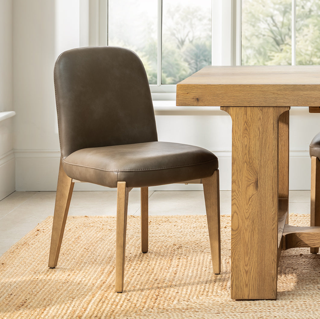 Smart and modern dining chair with a brown faux leather seat and wooden legs, positioned next to a wooden dining table in a bright room with a window and woven rug.