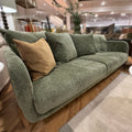 A three-seater sofa upholstered in textured sage green velvet, styled with four matching green pillows and one tan accent pillow, set in a modern living room environment.
