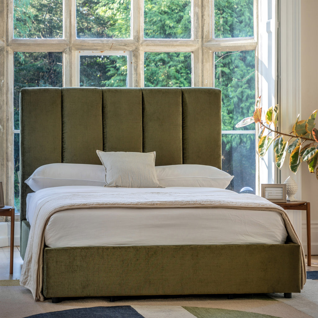 Green upholstered bed with white bedding in a room with large windows and plants.