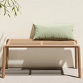 Modern doga outdoor bench with a light beige slatted frame and legs, styled with a sage green rectangular throw pillow, set against a textured off-white wall with leafy shadows.