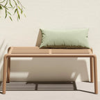 Modern doga outdoor bench with a light beige slatted frame and legs, styled with a sage green rectangular throw pillow, set against a textured off-white wall with leafy shadows.
