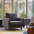 A modern gray upholstered armchair with a high back and tapered wooden legs, positioned in a living room setting with large windows showing trees outside.