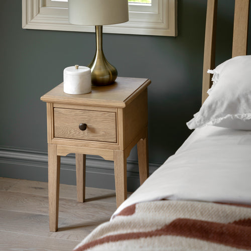 Wooden bedside table with lamp and candle next to a bed with white bedding and blanket.