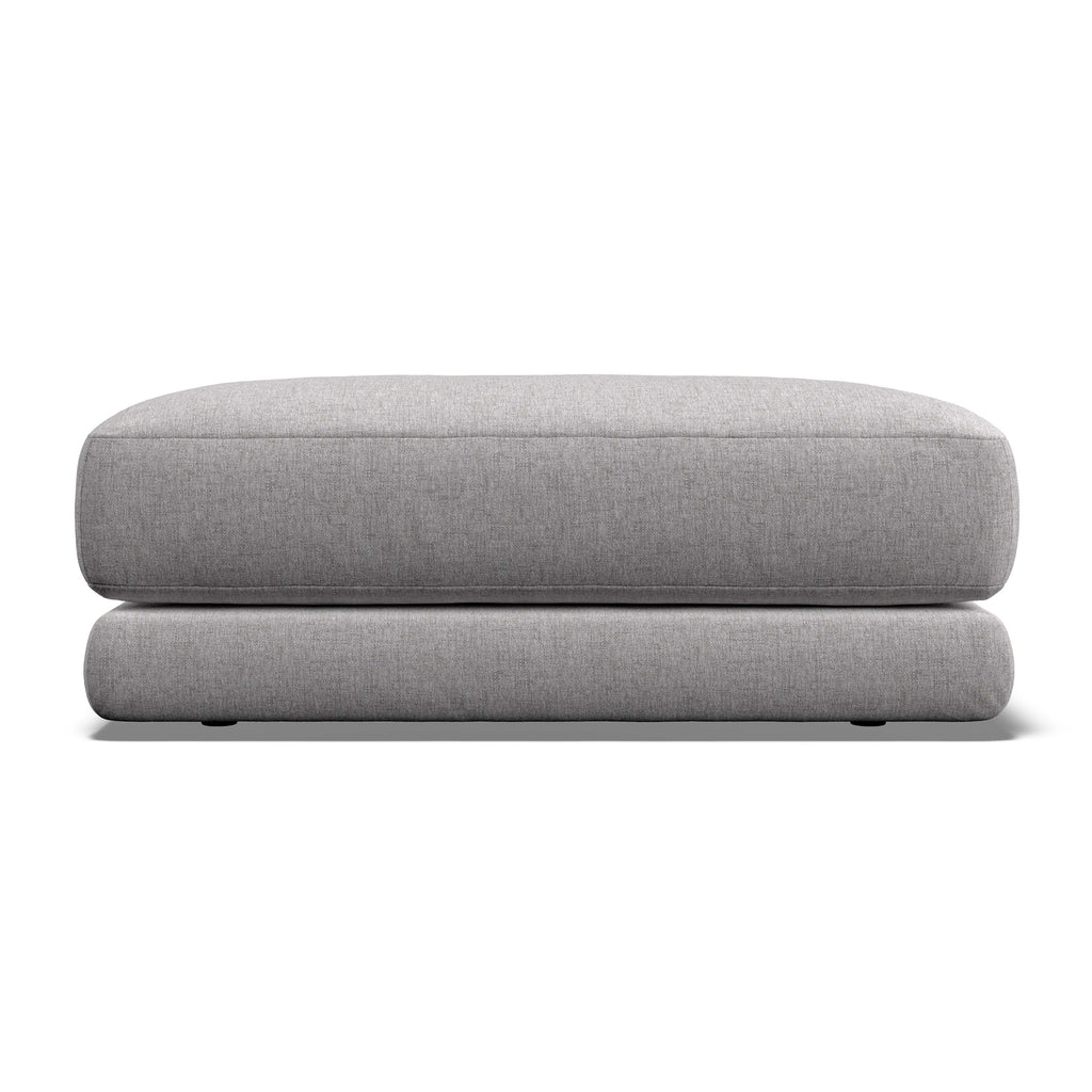A light gray upholstered ottoman or low stool with a thick rectangular seat cushion and rounded legs, shown against a plain white background.