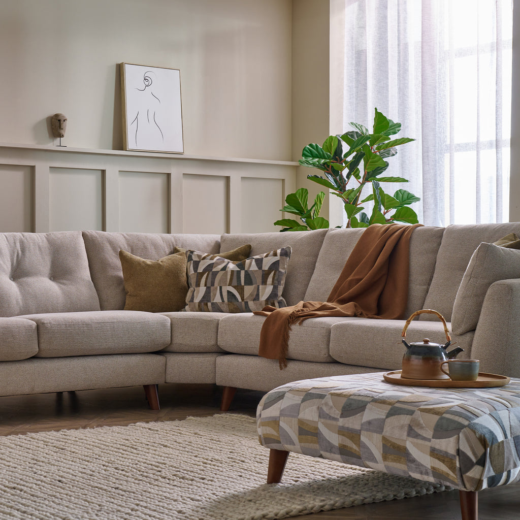 A modern beige sectional sofa in a styled living room, decorated with patterned and solid throw pillows and a brown throw blanket. A matching patterned ottoman sits in the foreground on a woven rug, with a window, curtains, and a green plant in the background.