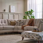 A modern beige sectional sofa in a styled living room, decorated with patterned and solid throw pillows and a brown throw blanket. A matching patterned ottoman sits in the foreground on a woven rug, with a window, curtains, and a green plant in the background.