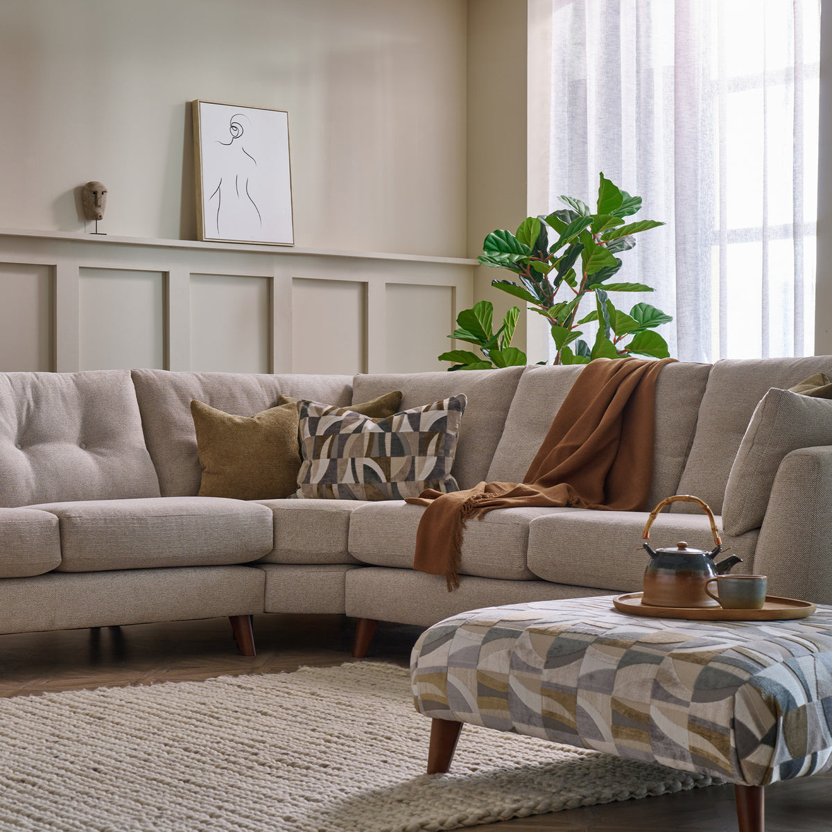 A modern beige sectional sofa in a styled living room, decorated with patterned and solid throw pillows and a brown throw blanket. A matching patterned ottoman sits in the foreground on a woven rug, with a window, curtains, and a green plant in the background.