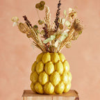 Decorative vase shaped with lemons filled, containing dried flowers on a wooden stand against a peach background.