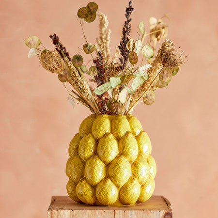 Decorative vase shaped with lemons filled, containing dried flowers on a wooden stand against a peach background.