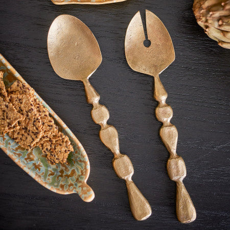 Two decorative metal brass salad servers on a dark surface with ceramic plates and food items