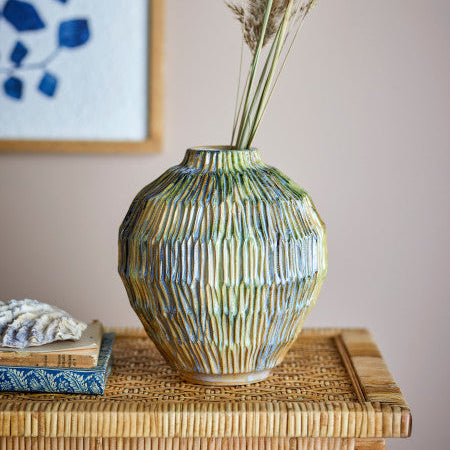 Decorative ceramic vase with textured surface on a wicker surface, with a framed abstract painting in the background.